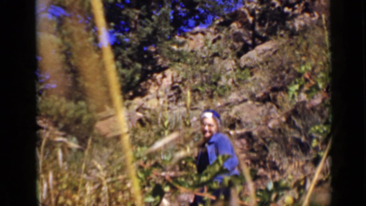 In 1939 Colorado home-movie footage, a woman in blue-purple clothing stands partly obscured by tall plants in a sunlit rural clearing.