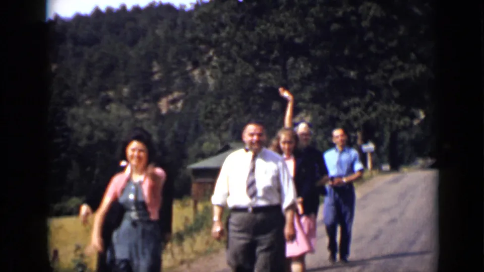 A woman in a dark dress or apron walks near the front as adults in dress clothes continue toward the camera on the sunlit unpaved road in Colorado.