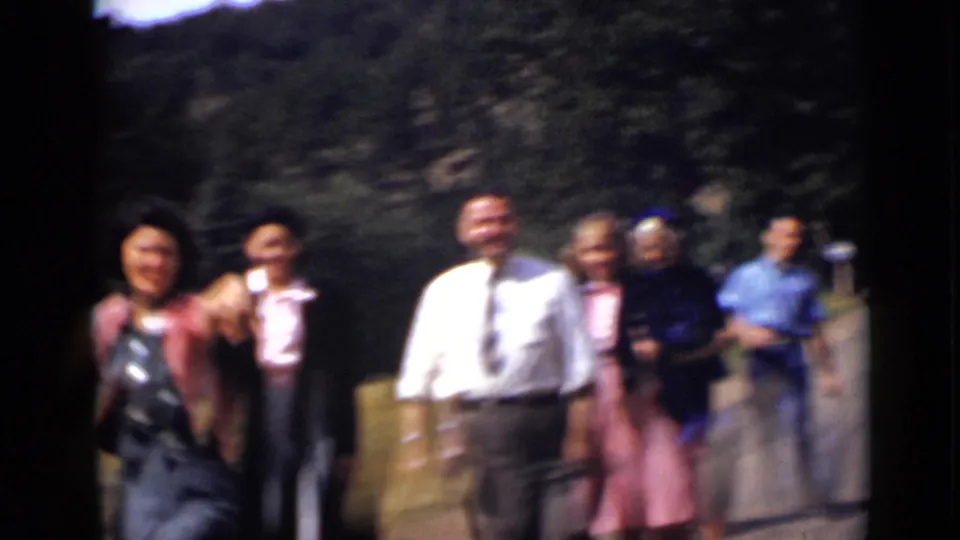 The group reaches a closer, blurrier point in the 8mm frame, with smiling adults in dress clothes spread across the wooded roadside scene.