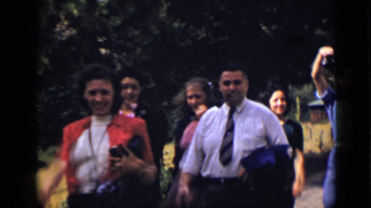 Adults walk outdoors near trees, including a woman in red and a man in a white shirt and tie.