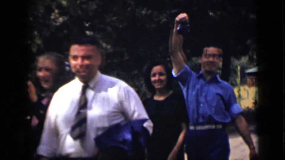 Adults in 1939 Colorado walk through a shaded outdoor area as a man in blue raises a camera and others smile nearby.