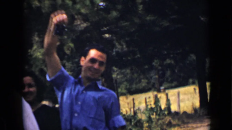 A smiling man in a blue shirt raises a small camera in 1939 Colorado home-movie footage, with trees and another adult partly visible.