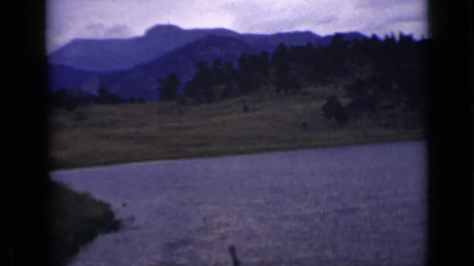1939 Colorado home-movie frame showing water, open banks, evergreen trees, and hazy mountains beyond.