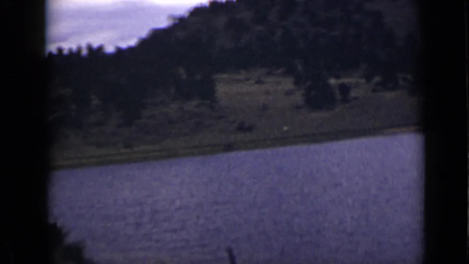 Soft 8mm frame of a Colorado waterway with a grassy edge, dark hillside trees, and pale sky.