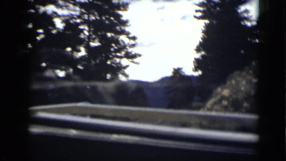 Roadside view from a vehicle in Colorado, with a railing or car edge, trees, and distant hillside under bright sky.
