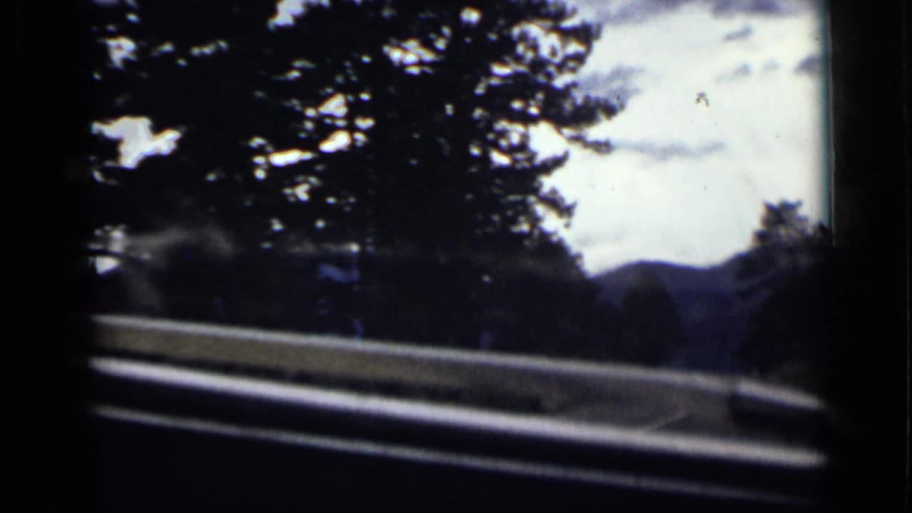 Colorado mountain road view from inside a moving vehicle, with guardrail, evergreens, and hazy hills visible through the windshield.