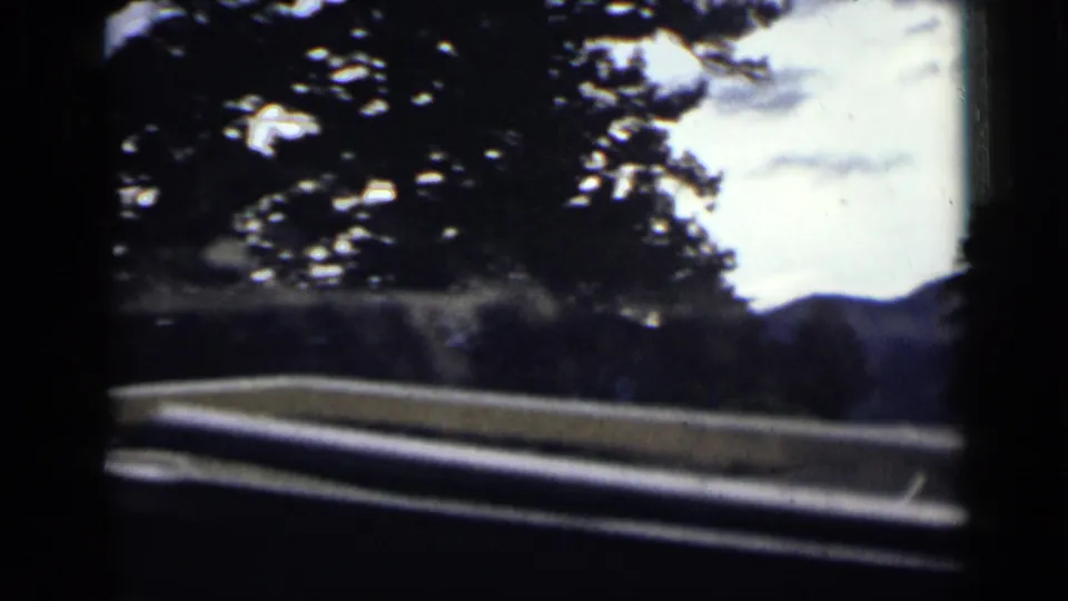 8mm home-movie frame from Colorado showing a blurred in-car view of trees, guardrail, and a mountain ridge beyond the road.