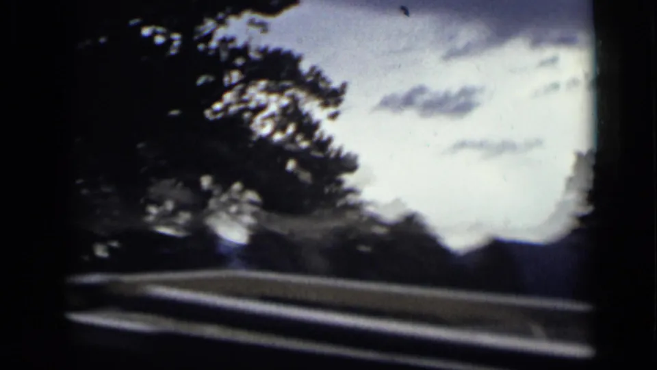 Final 1939 Colorado road-travel frame with silhouetted evergreens, pale clouds, guardrail, and distant mountain forms through the windshield.