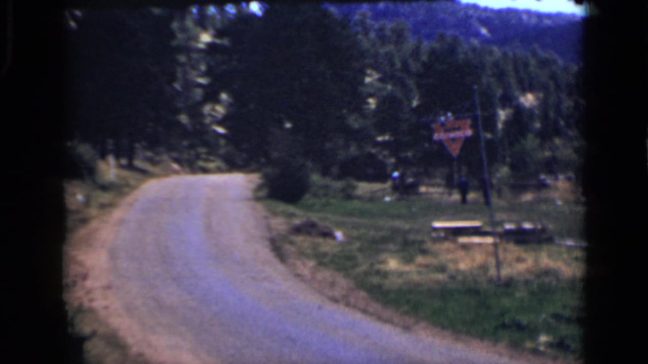 Colorado 1939 8mm view down a winding roadside with trees, a grassy picnic area, benches, and a red Conoco sign near the right edge.