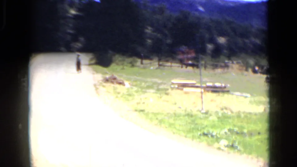 Brightly exposed Colorado 1939 home-movie frame showing a person walking along the road near grass, trees, picnic tables, and a roadside sign.