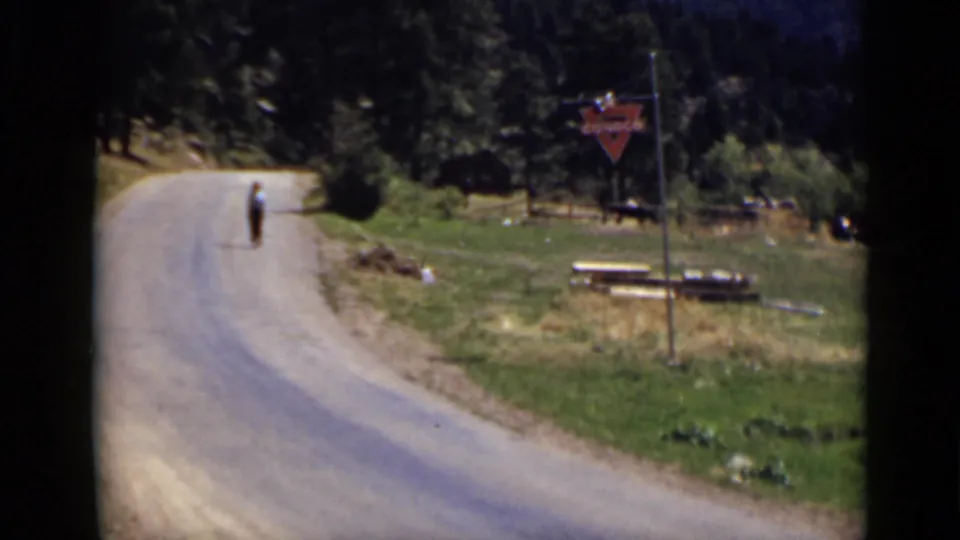 Colorado 1939 8mm frame of a person walking on a curving mountain road, with trees, picnic tables, roadside grass, and a red Conoco sign visible.
