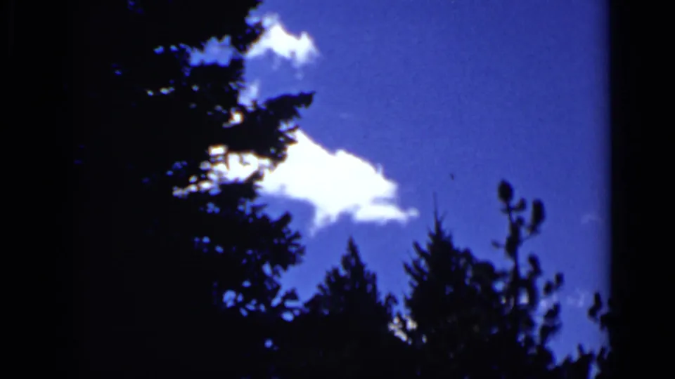 1939 Colorado home-movie frame with white clouds against saturated blue sky, bordered by dark conifer shapes.
