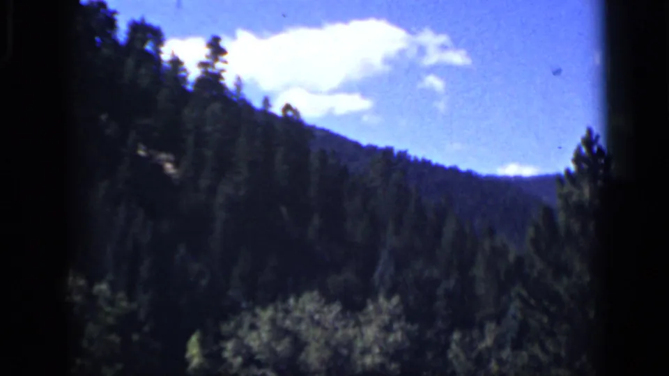 1939 Colorado home-movie frame showing a wooded mountain slope, meadow foreground, and white clouds in a blue sky.