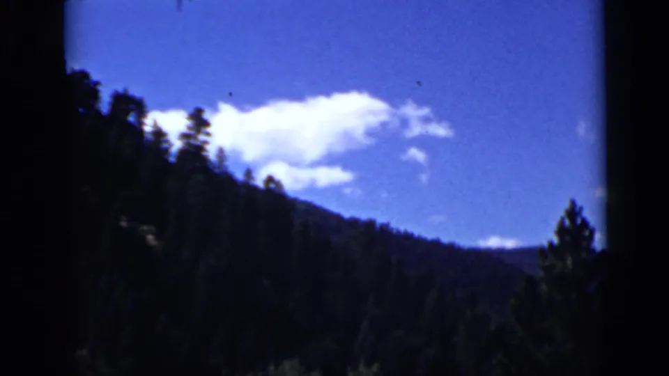 Colorado 8mm frame of a dark forested ridge with treetops silhouetted against blue sky and white clouds.
