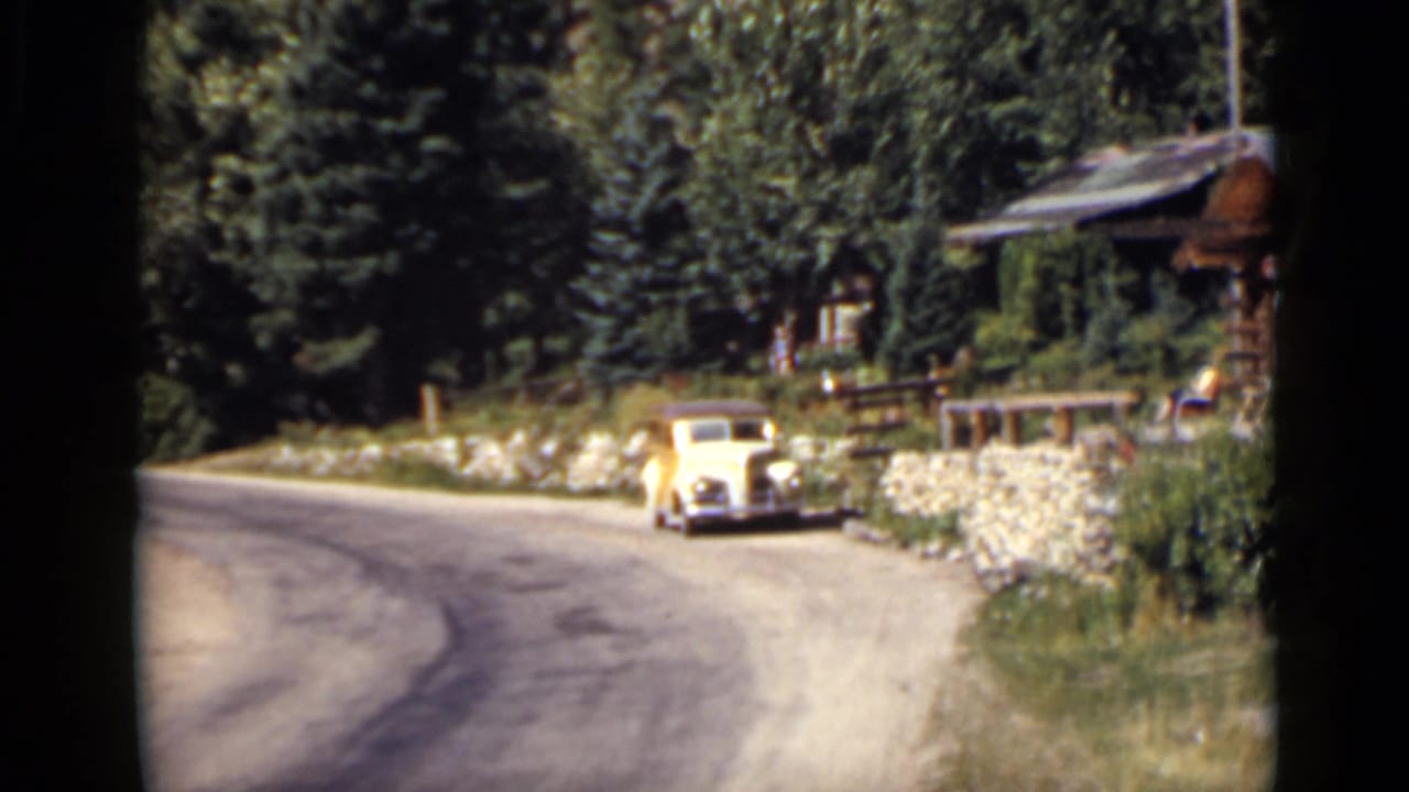 Vintage pale car parked beside a curved road near a log cabin and trees.