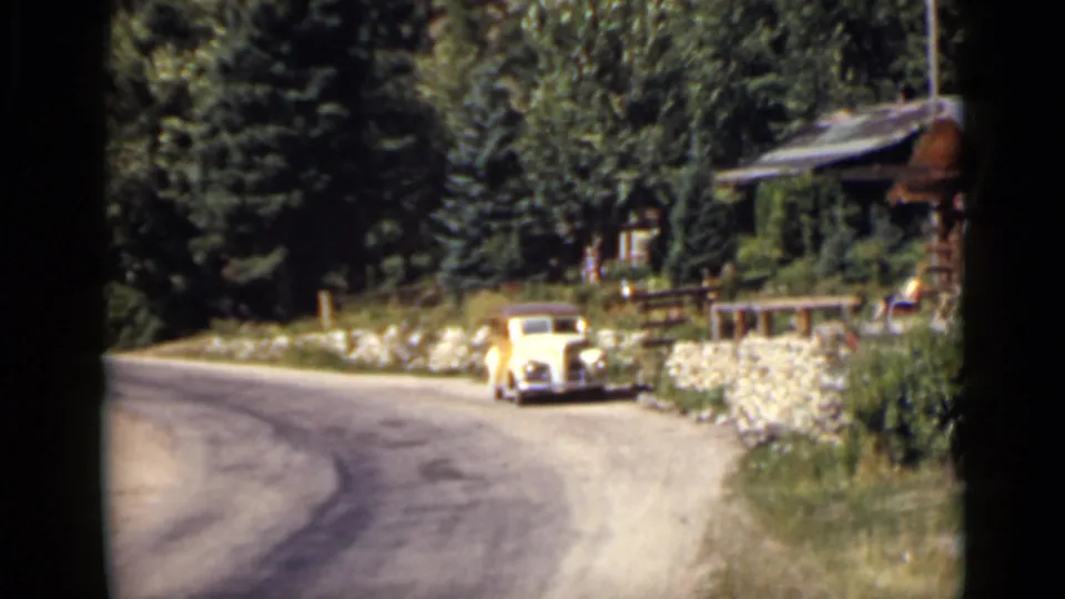 1939 Colorado 8mm frame showing a pale vintage car beside a curved road near a log cabin, stone wall, and evergreens.