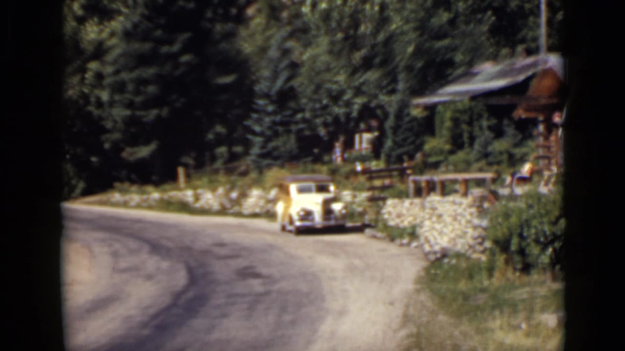Vintage car near a roadside cabin with trees and a low stone wall.