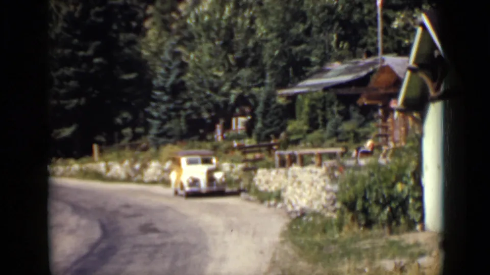 1939 Colorado 8mm frame with a pale vintage car near a wooded cabin scene, stone wall, road curve, and foreground building edge.