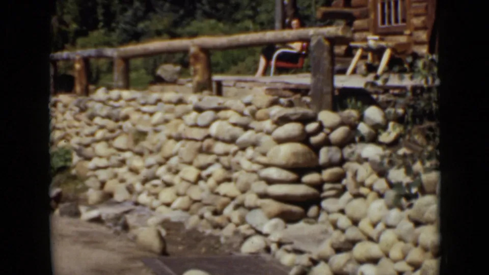 1939 Colorado cabin scene with a stacked stone wall, wooden porch railing, trees, and a partial light-colored automobile.