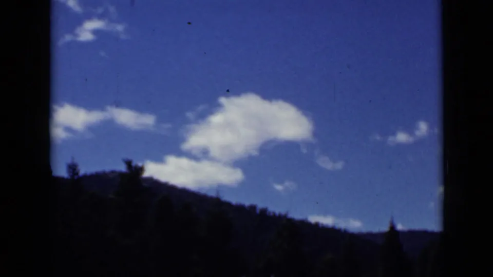 1939 Colorado home-movie view of white clouds in a blue sky over a dark forested mountain ridge.