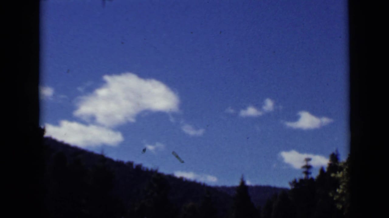 Blue sky with white clouds above dark wooded hills, framed by black film or window edges.