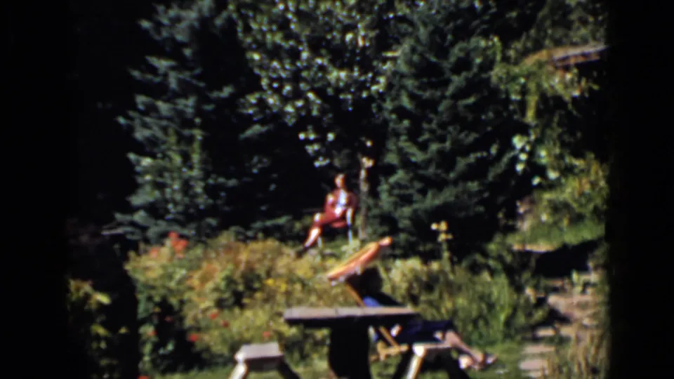 1939 Colorado home-movie garden view with a distant woman in red seated among foliage near a picnic table.