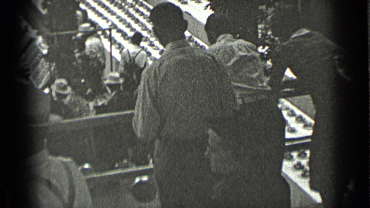 1939 New York 8mm frame showing men in coats and hats near railings, with a crowded public hall visible beyond them.
