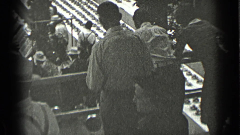 1939 New York 8mm frame showing men in coats and hats near railings, with a crowded public hall visible beyond them.