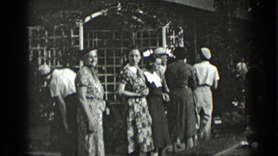 Women in late-1930s-style dresses and hats stand outside a New York building with a latticed archway while other adults move nearby.