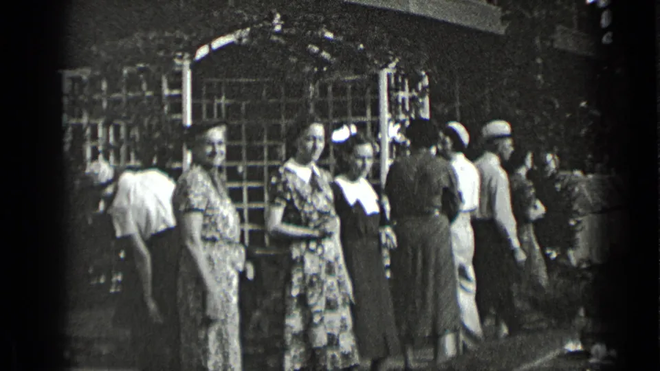 A New York 1939 home-movie frame shows women in patterned dresses near a building entrance, with men and women passing in the background.
