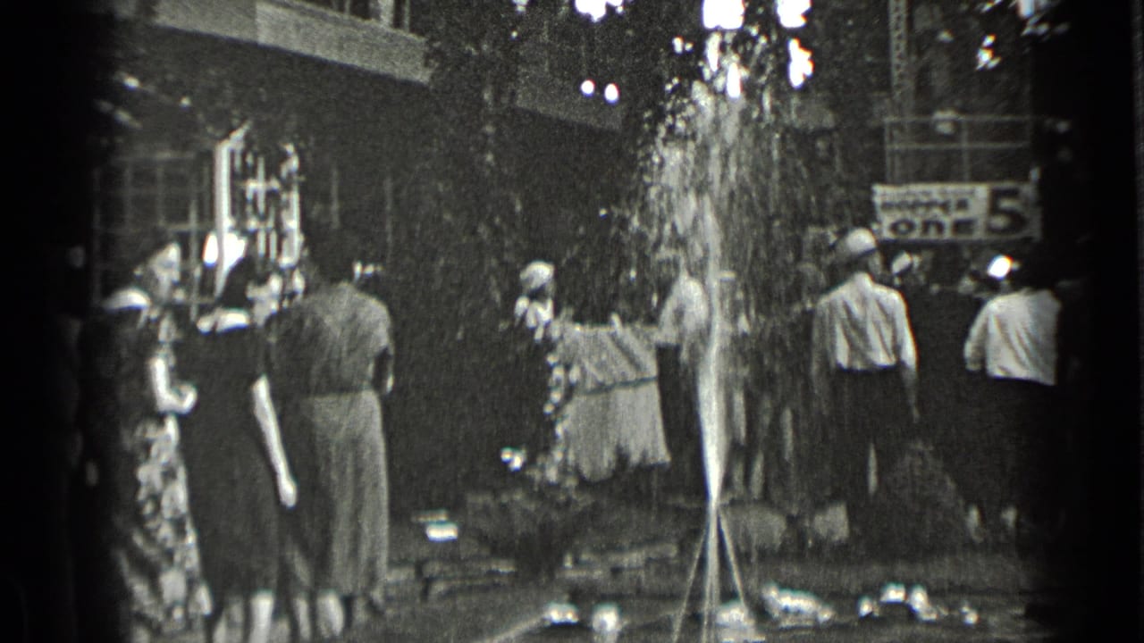 Adults in 1939 New York public space watch water spray from a fountain, with trees, a building edge, and a partial sign in the background.