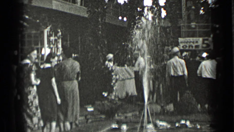 Adults in 1939 New York public space watch water spray from a fountain, with trees, a building edge, and a partial sign in the background.