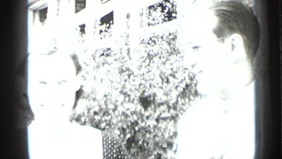 Bright black-and-white home-movie frame of two adults outdoors in New York, standing near foliage and a building.