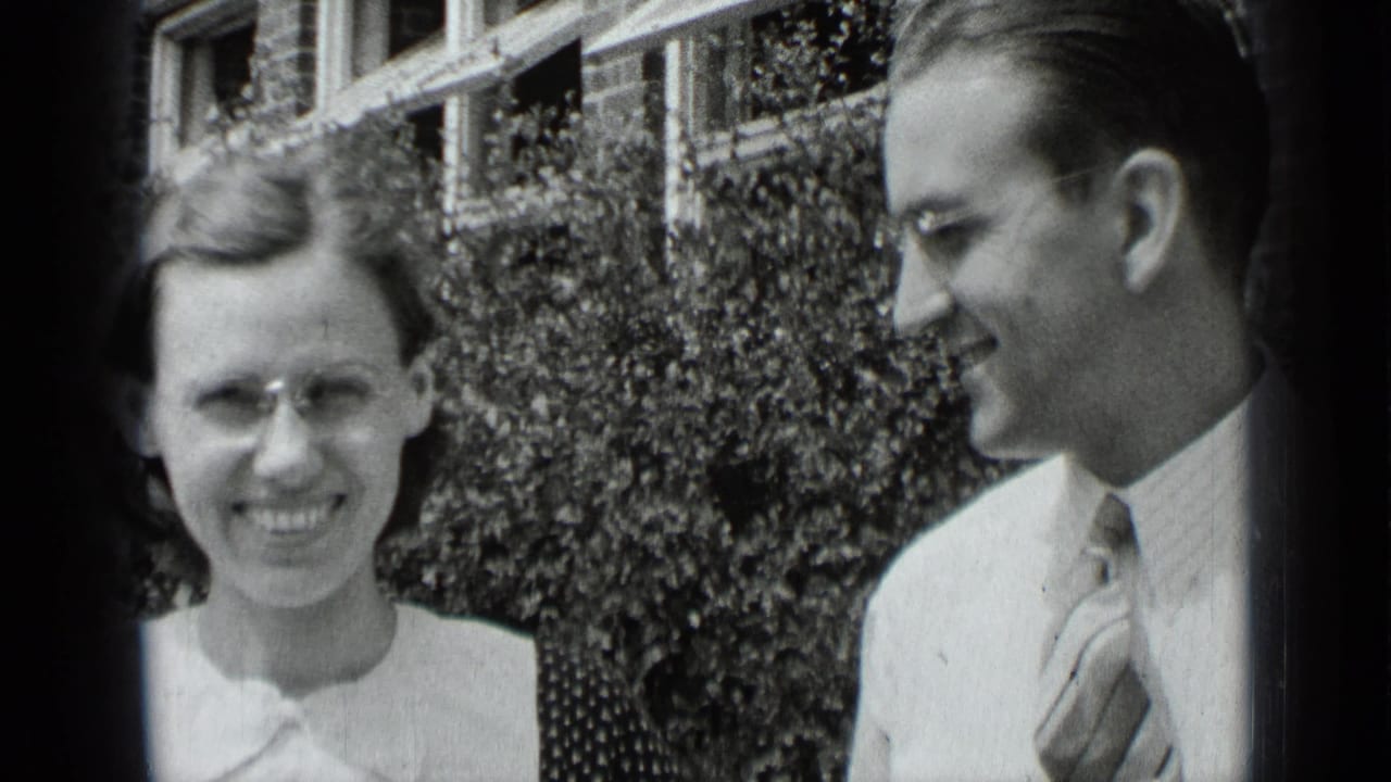 Woman and man smiling outdoors in front of hedges and a brick building.