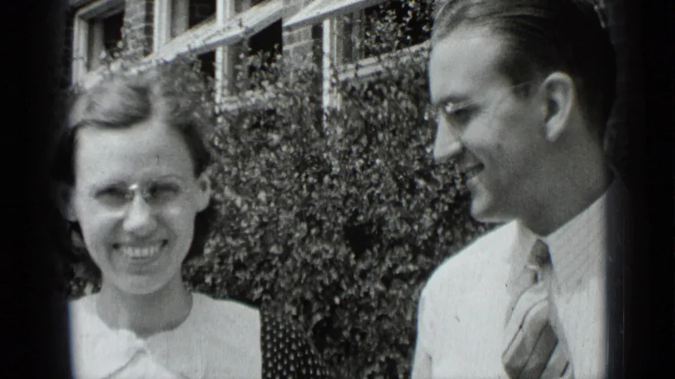 In 1939 New York home-movie footage, a woman with glasses and a man in a tie smile outdoors before hedges and brick windows.