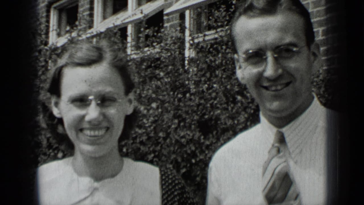 The couple stands outside in New York, both wearing glasses, with the woman smiling directly and the man looking toward the camera.