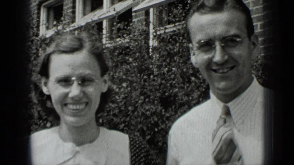 The couple stands outside in New York, both wearing glasses, with the woman smiling directly and the man looking toward the camera.