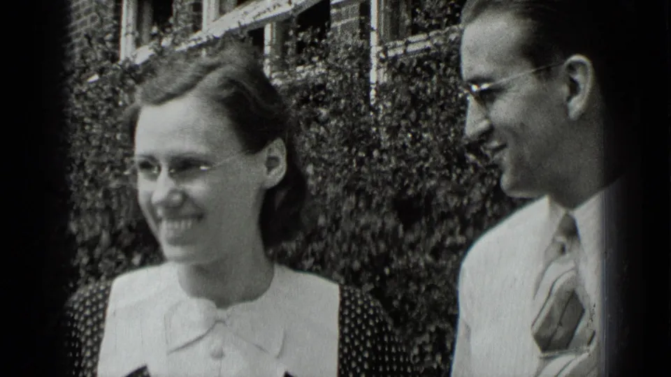 In the 1939 outdoor home movie, the woman smiles off to the side while the man turns toward her near the hedge-lined brick building.