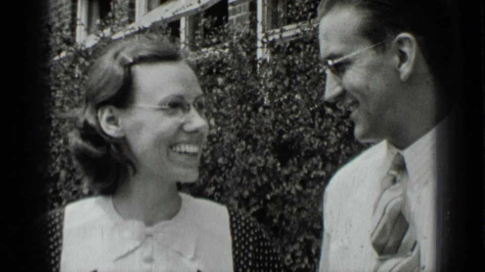The woman appears to laugh or smile broadly toward the man in this 1939 New York 8mm frame, with hedges and a brick facade behind them.