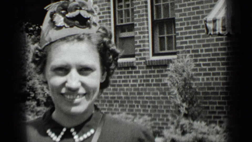 In 1939 New York home-movie footage, a woman in a decorative hat and dark dress smiles near a brick building and shrubs.