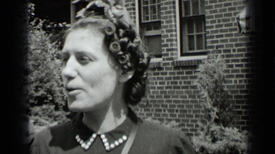 The woman appears in side view, speaking or reacting while standing outside a brick building in the 1939 New York home movie.