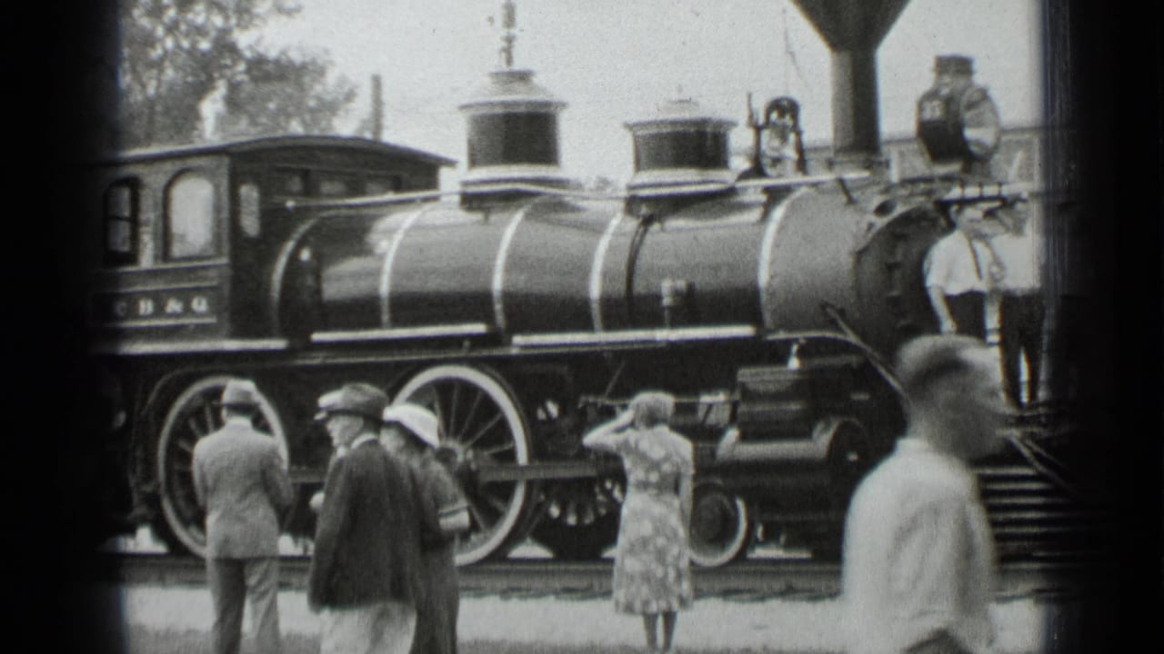 Townsfolk stand beside a black steam locomotive with large wheels and cab lettering.