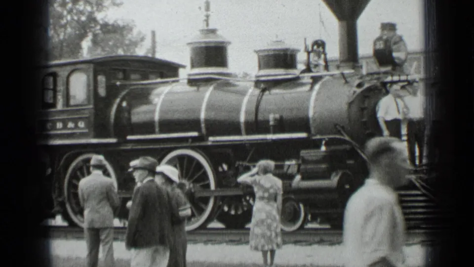 1939 New York home-movie frame showing visitors in period clothing beside a black steam locomotive on display.
