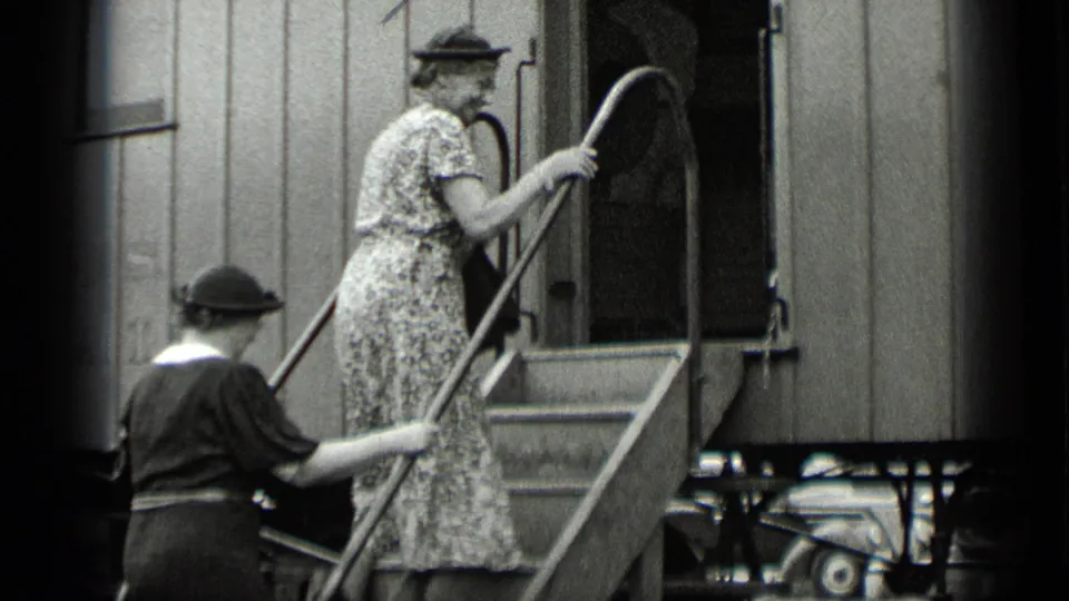 A woman in a patterned dress and hat climbs the railcar stairs as another passenger waits below.