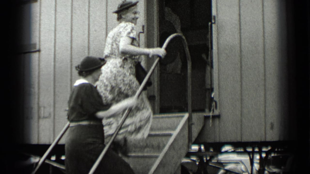 Women in hats climb exterior steps into a rail car as another passenger stands nearby.