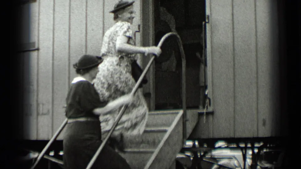 In 1939 New York home-movie footage, women in period dress and hats board a rail car using exterior steps and curved handrails.