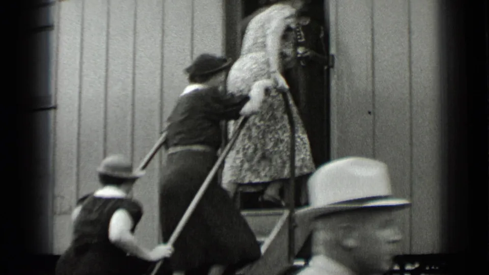 Women in dresses and hats move up the steps into a rail car, with a passing foreground passenger partly obscuring the view.