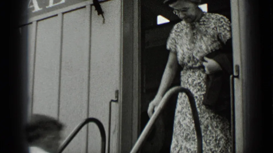 A woman in a floral dress and hat appears at the rail-car doorway, holding the handrail in black-and-white 8mm footage.