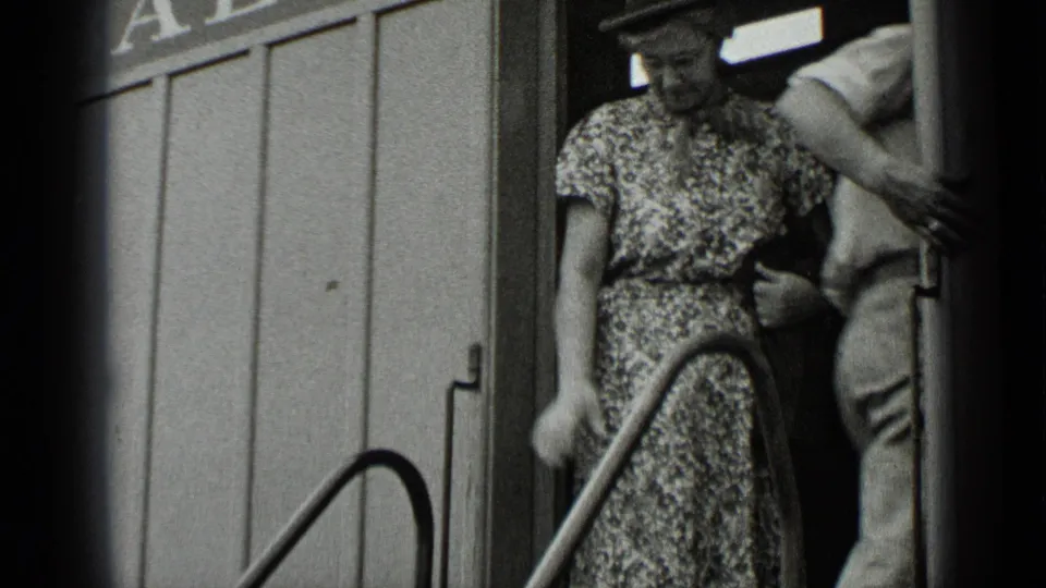 The woman in the floral dress is seen on the rail-car steps beside another passenger, framed tightly by the doorway and handrail.
