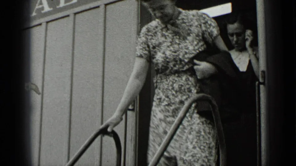 In 1939 New York fair footage, a woman in a patterned dress descends metal ride stairs with another adult visible in the doorway.